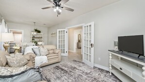 Un salon avec un canapé beige, un téléviseur posé sur un meuble blanc et un ventilateur de plafond.