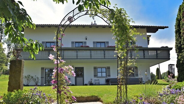 Un bâtiment à deux étages doté d’un balcon, entouré d’un jardin bien entretenu, agrémenté de plantes en fleurs et d’un chemin de pierre menant à l’entrée.