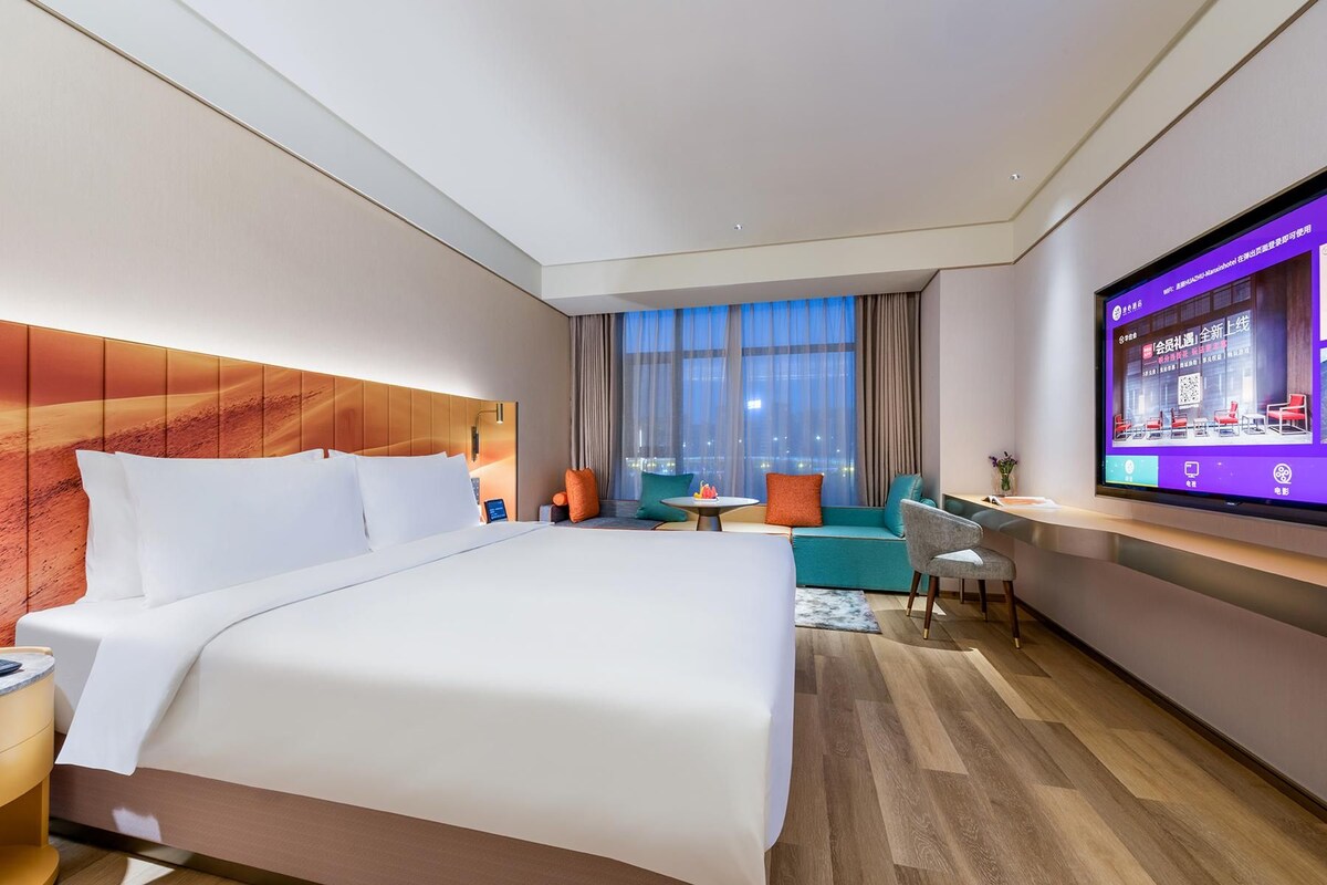 A hotel room with a large bed, a seating area with a table, a TV on the wall, and a view of the city.