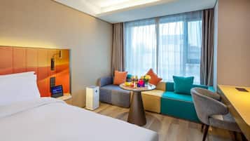 A hotel room with a bed, a sofa, a small table with fruits and drinks, and a desk.