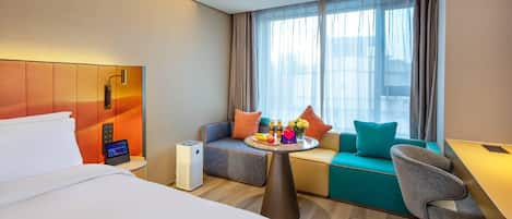 Quarto de hotel com cama, sofá, pequena mesa com frutas e bebidas, e escrivaninha.