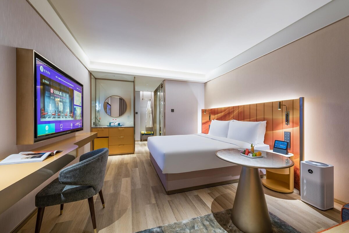 A modern hotel room with a bed, bedside table, TV, desk, chair, and a small round table with a tablet and a speaker.