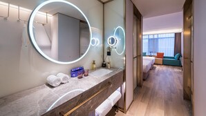 Un baño de hotel moderno con un espejo grande, luces redondas, una encimera de mármol y una cama visible en la habitación contigua.
