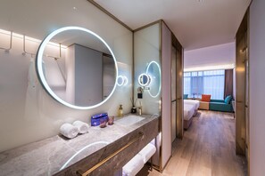 Un baño de hotel moderno con un espejo grande, luces redondas, una encimera de mármol y una cama visible en la habitación contigua.