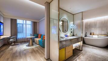 A modern hotel room with a large window, a sofa, a round table, a TV, a bathroom with a bathtub, and a vanity with a mirror.