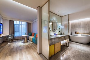 Una habitación de hotel moderna con un ventanal grande, un sofá, una mesa redonda, un televisor, un baño con bañera y un tocador con espejo.