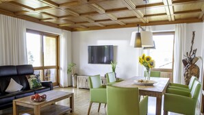 Un salon moderne avec un plafond en bois, un canapé noir, des fauteuils verts, une table basse en bois et une table à manger agrémentée d’un vase de tournesols.