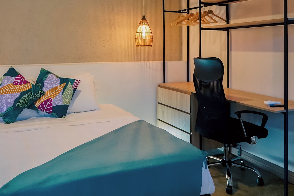 A bedroom with a bed, a desk, a chair, a hanging light fixture, and an air conditioning unit.