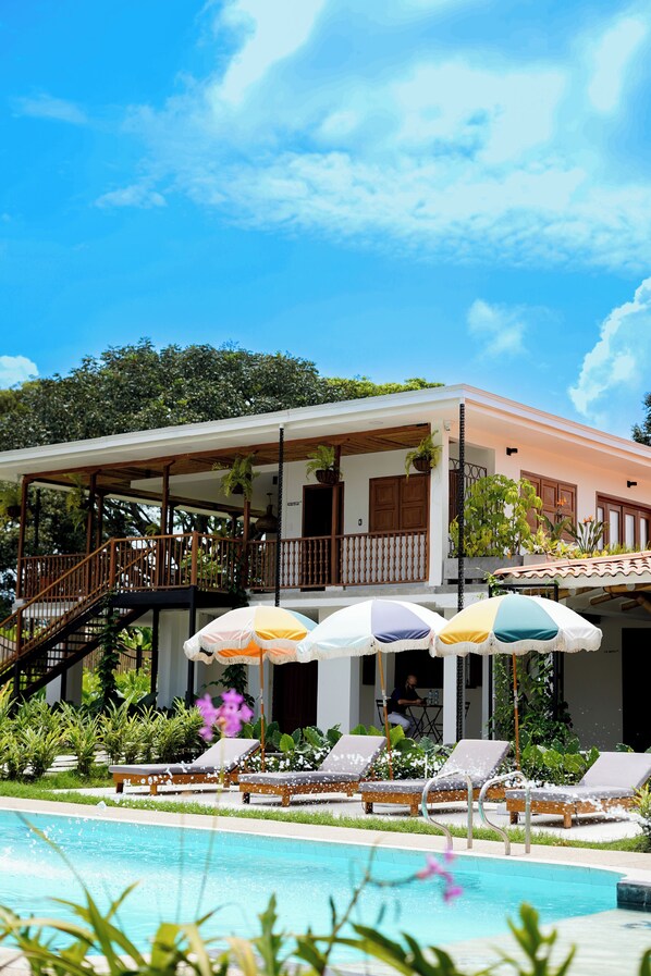 Zona junto a la piscina con sillones de descanso y sombrillas, una escalera que conduce a una terraza cubierta y exuberante vegetación al fondo.