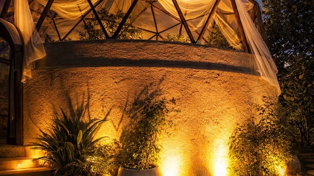 Une structure en forme de dôme géodésique recouverte d’une toile semi-transparente, entourée de plantes en pot et éclairée par un éclairage d’ambiance.