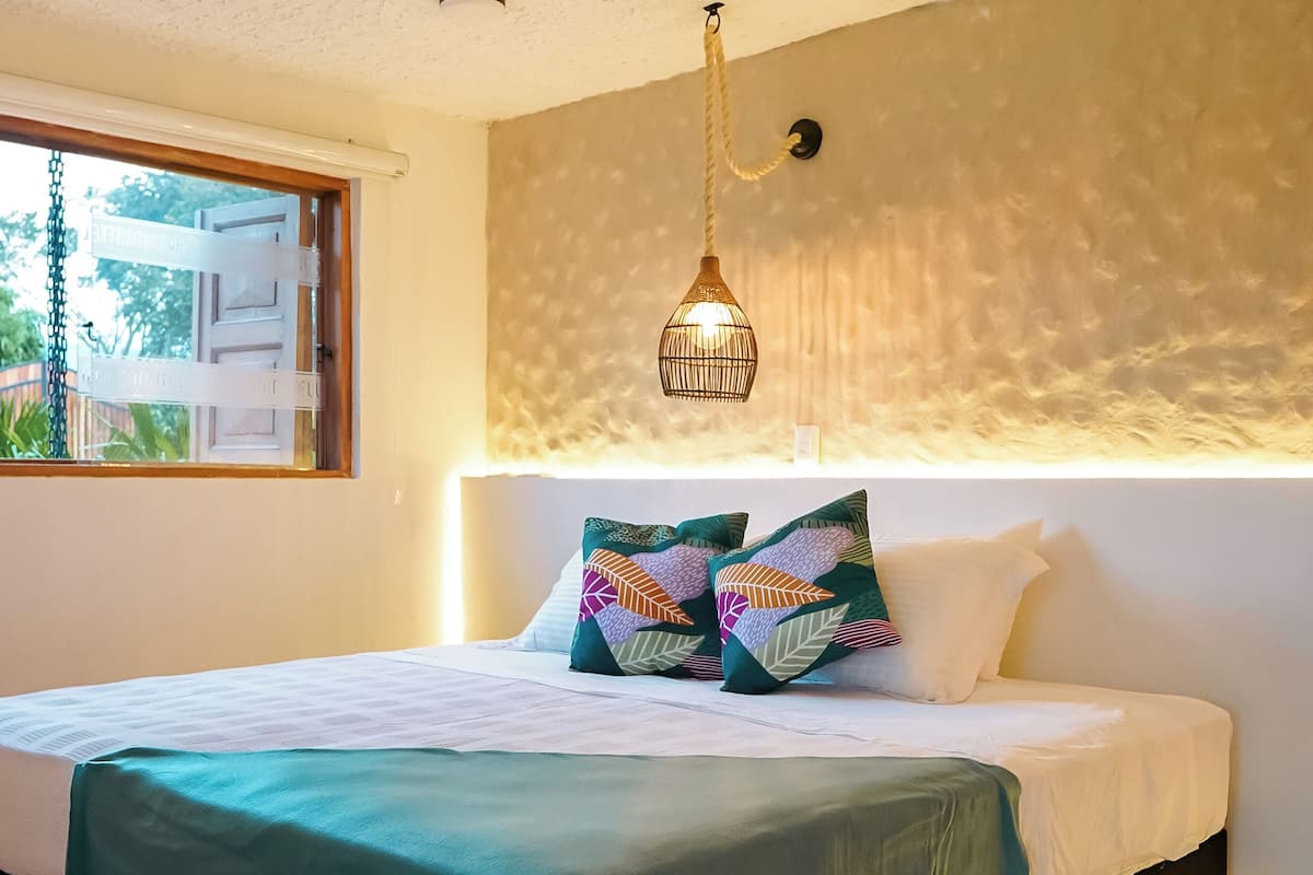Ett sovrum med en säng, kuddar, en hängande lampa och ett fönster med persienner.