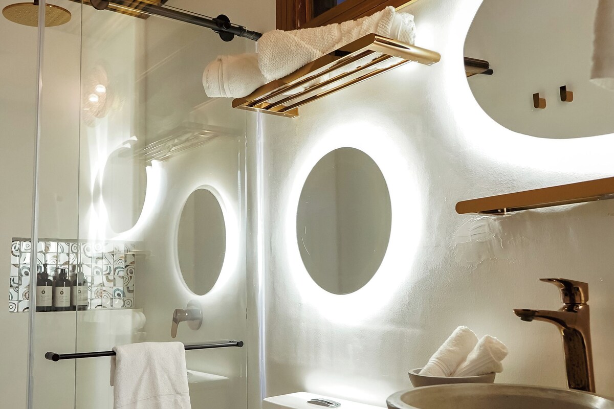 A modern bathroom with a glass-enclosed shower, a toilet, a sink with a bowl, and a mirror with circular lighting.