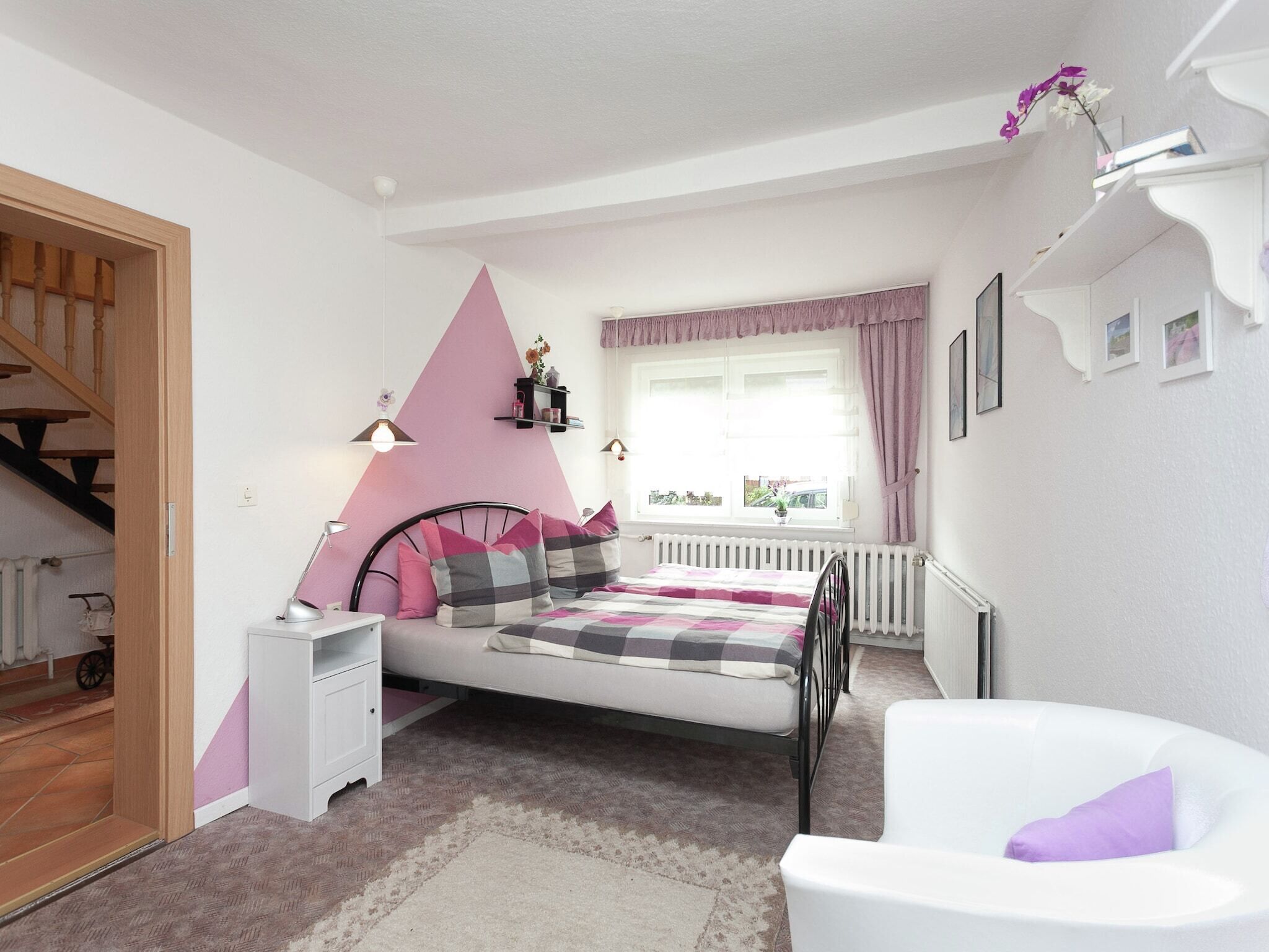 A bedroom with a bed, a white chair, a radiator, a window with curtains, and a staircase.
