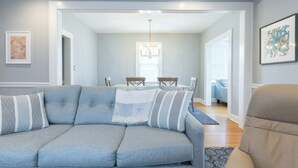 Una sala de estar moderna con un sofá gris, un comedor con una araña, y un pasillo que conduce a otra habitación.