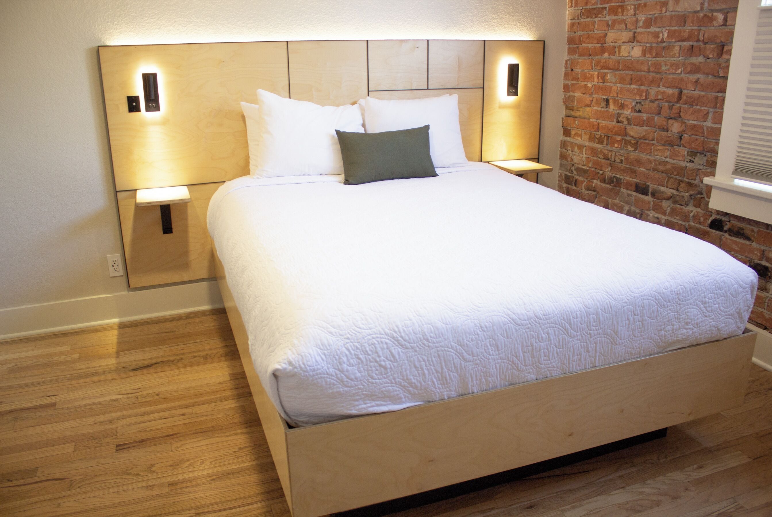 A neatly made bed with a wooden headboard, two bedside lamps, and a brick wall to one side.