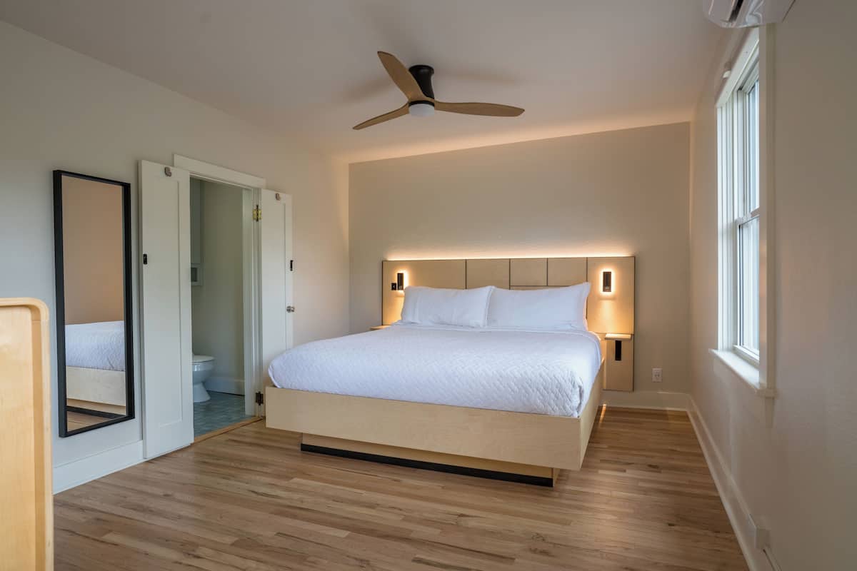 A bedroom with a bed, a ceiling fan, bedside lamps, a mirror, and a window.