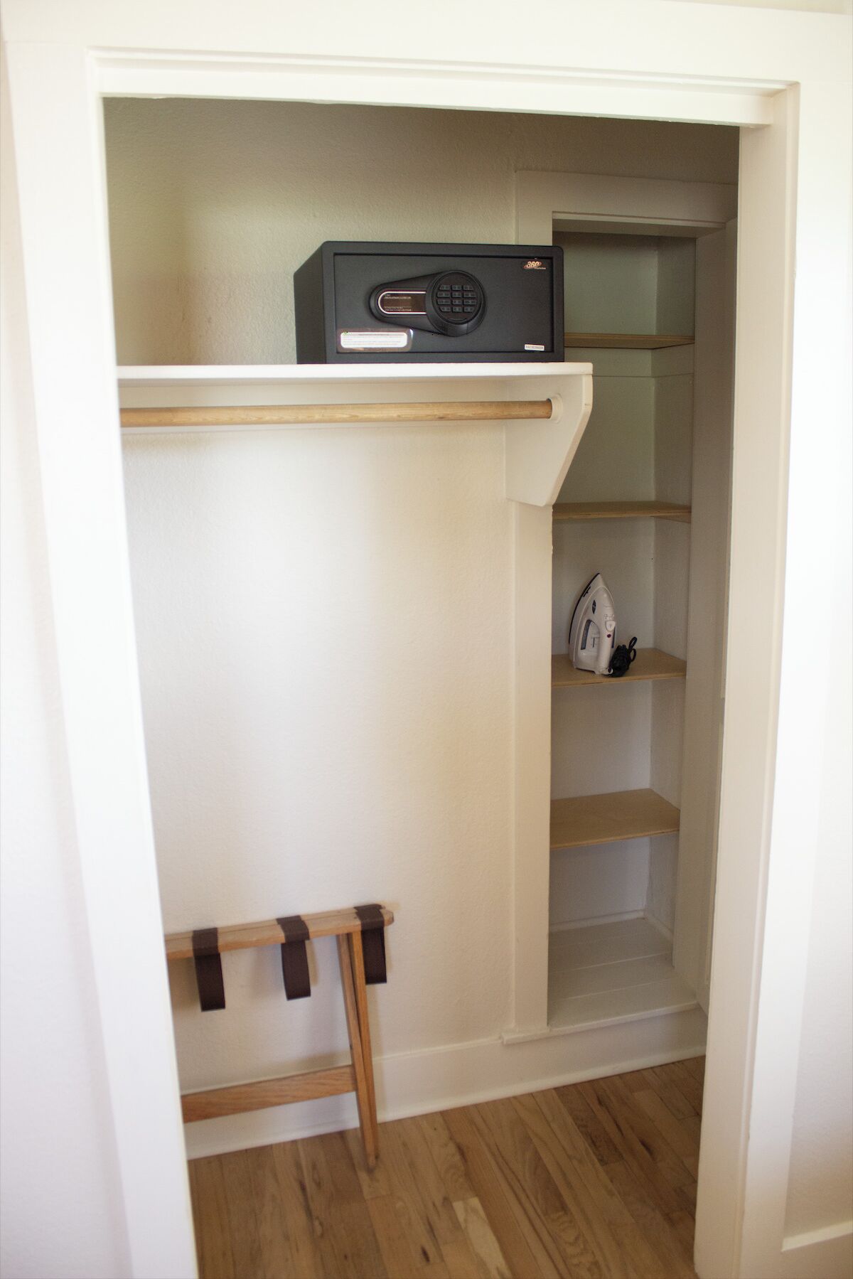 A small, enclosed closet with a wooden shelf and a hanging rod, containing a few items including a white iron and a black appliance.