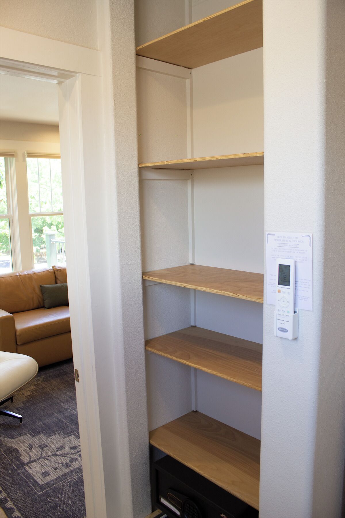 A room with a built-in wooden shelving unit against the wall, a thermostat mounted on the wall, and a view of a living area with a sofa and a window.