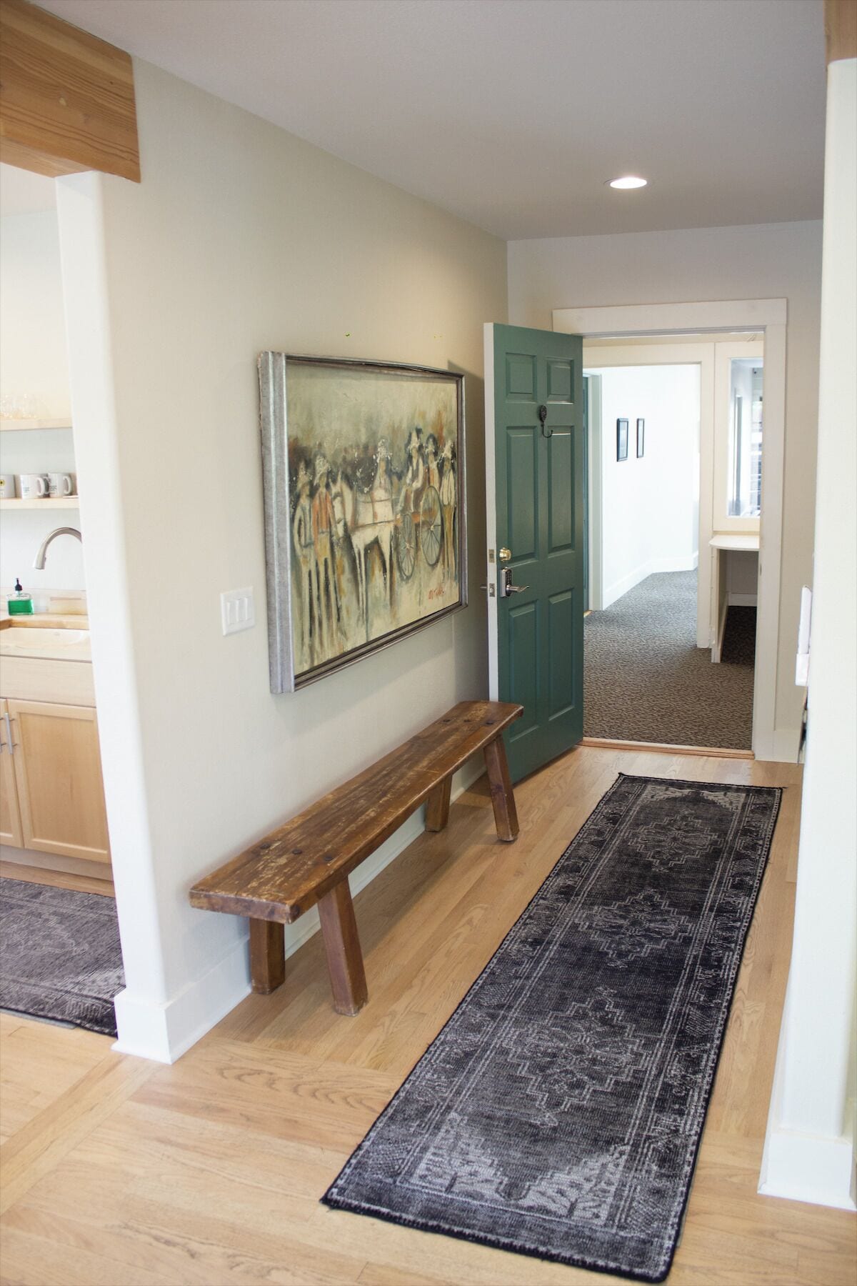 A hallway with a wooden bench, a dark patterned rug, a green door, and a painting on the wall.