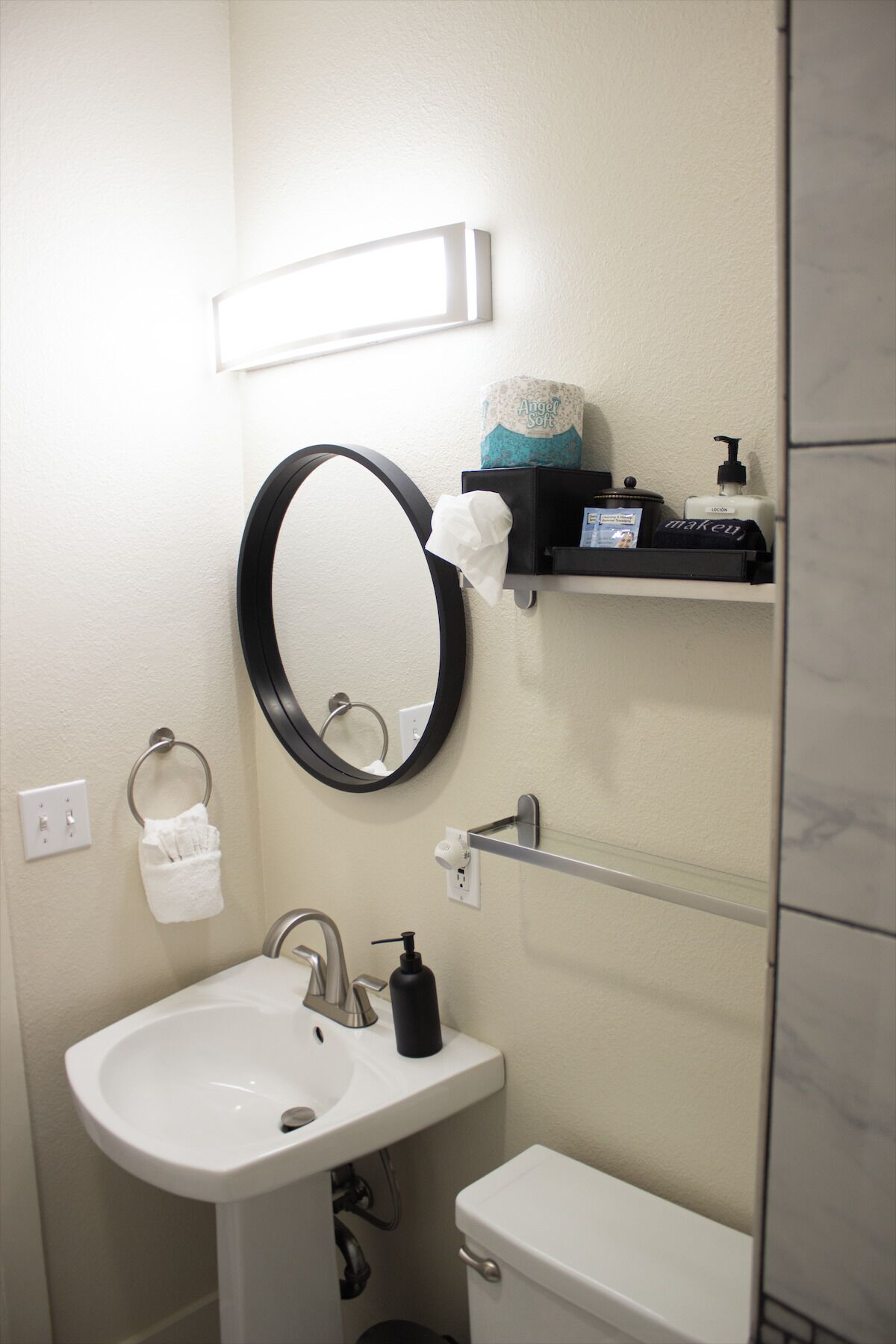 Une salle de bain avec un lavabo blanc, des toilettes, un miroir rond et une étagère contenant des articles de toilette.