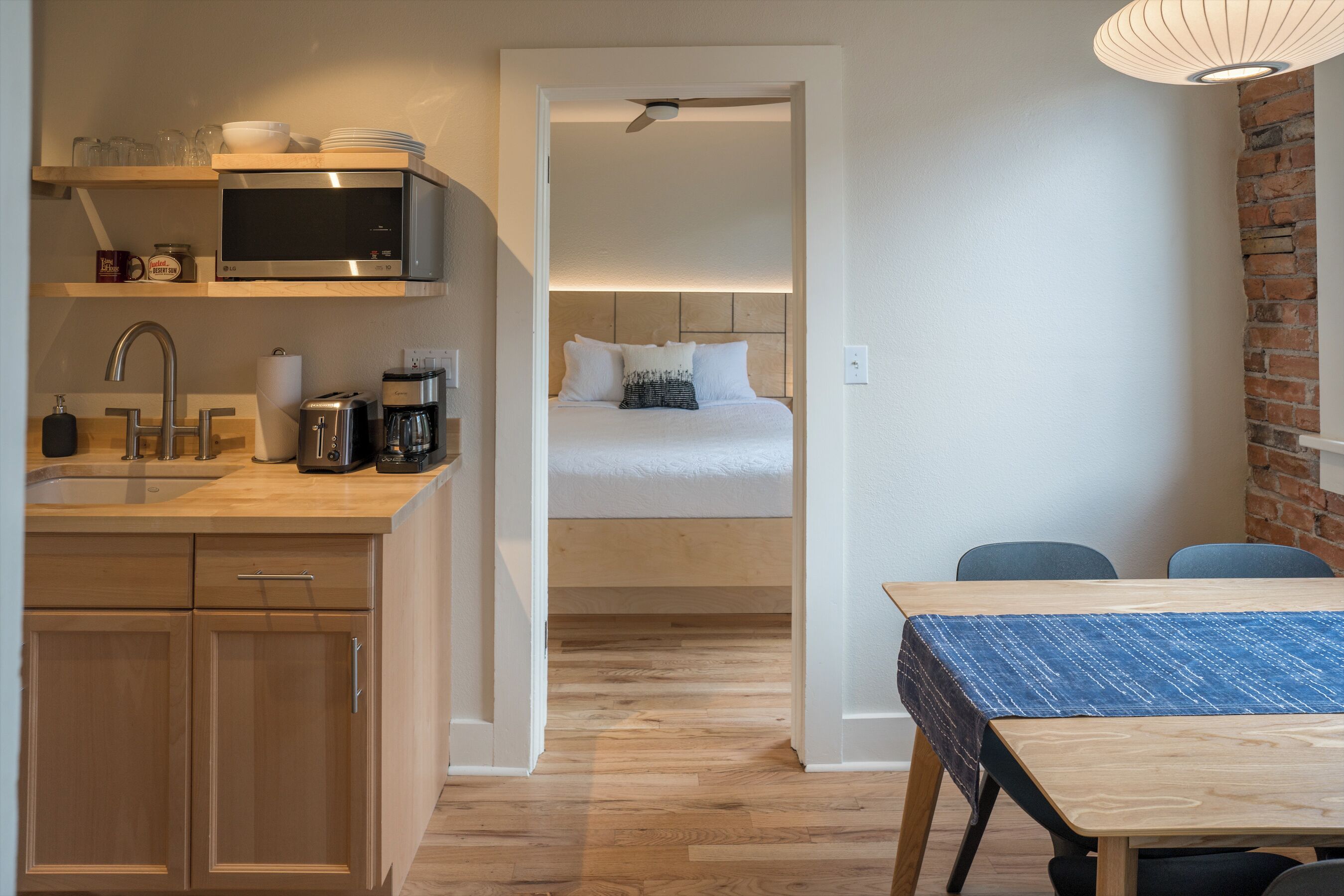 A compact hotel room with a kitchenette, a bed with white bedding, a dining table, and a brick accent wall.