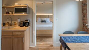 A compact hotel room with a kitchenette, a bed with white bedding, a dining table, and a brick accent wall.