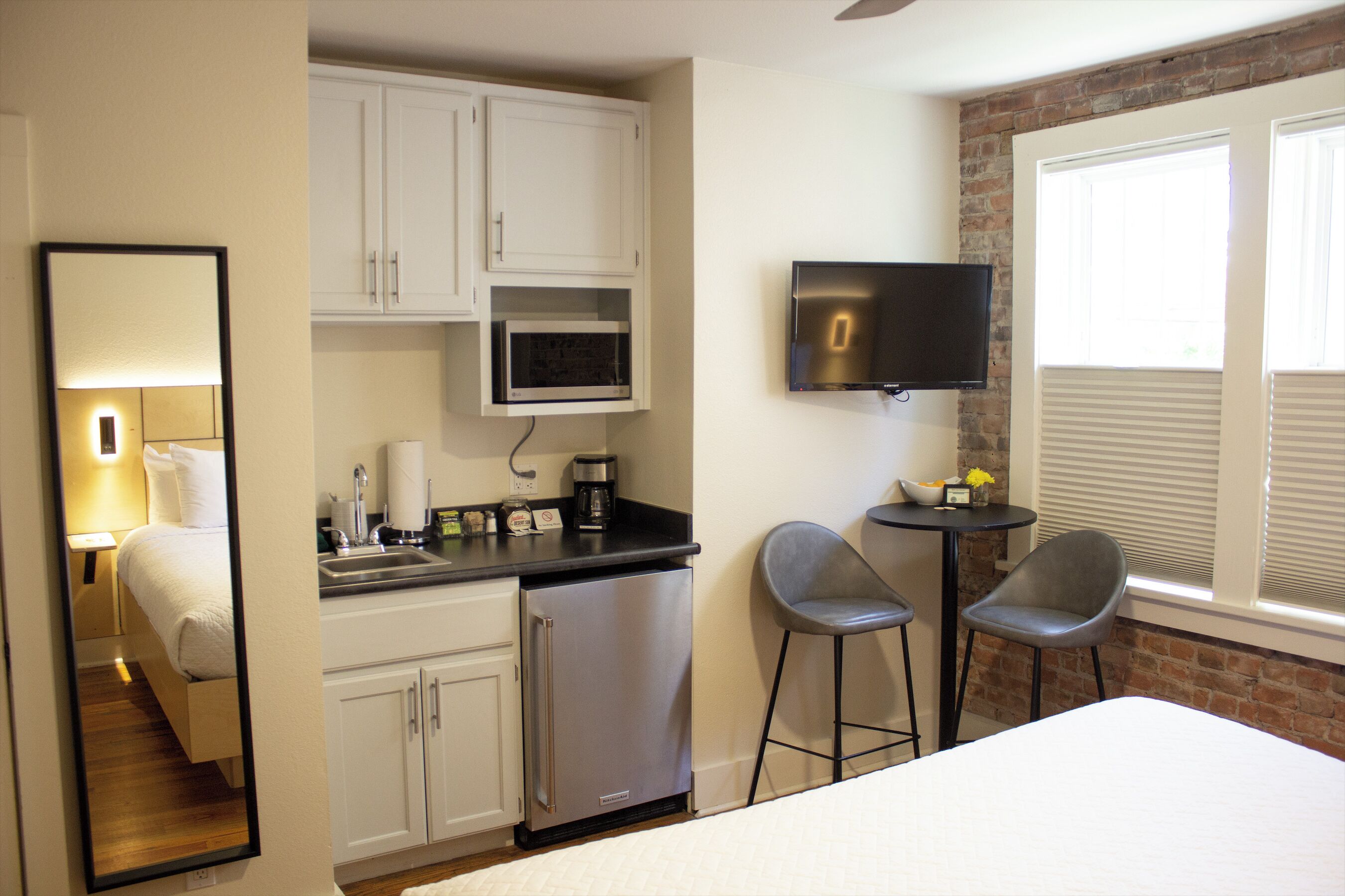 Habitación de hotel compacta con cama, kitchenette con microondas, refrigerador pequeño, televisor montado en la pared y ventana con persianas.