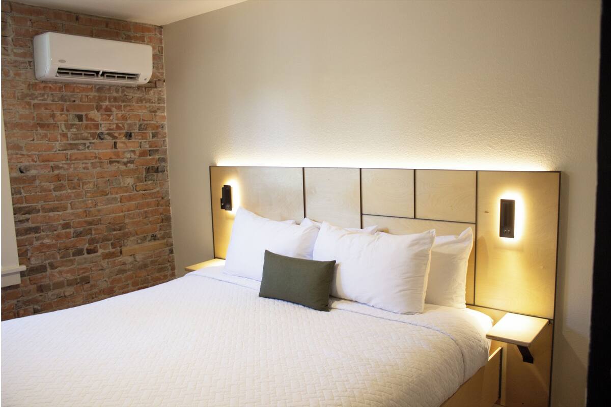 A neatly made bed with white bedding and a single green pillow, a brick wall, and a modern headboard with built-in lighting.