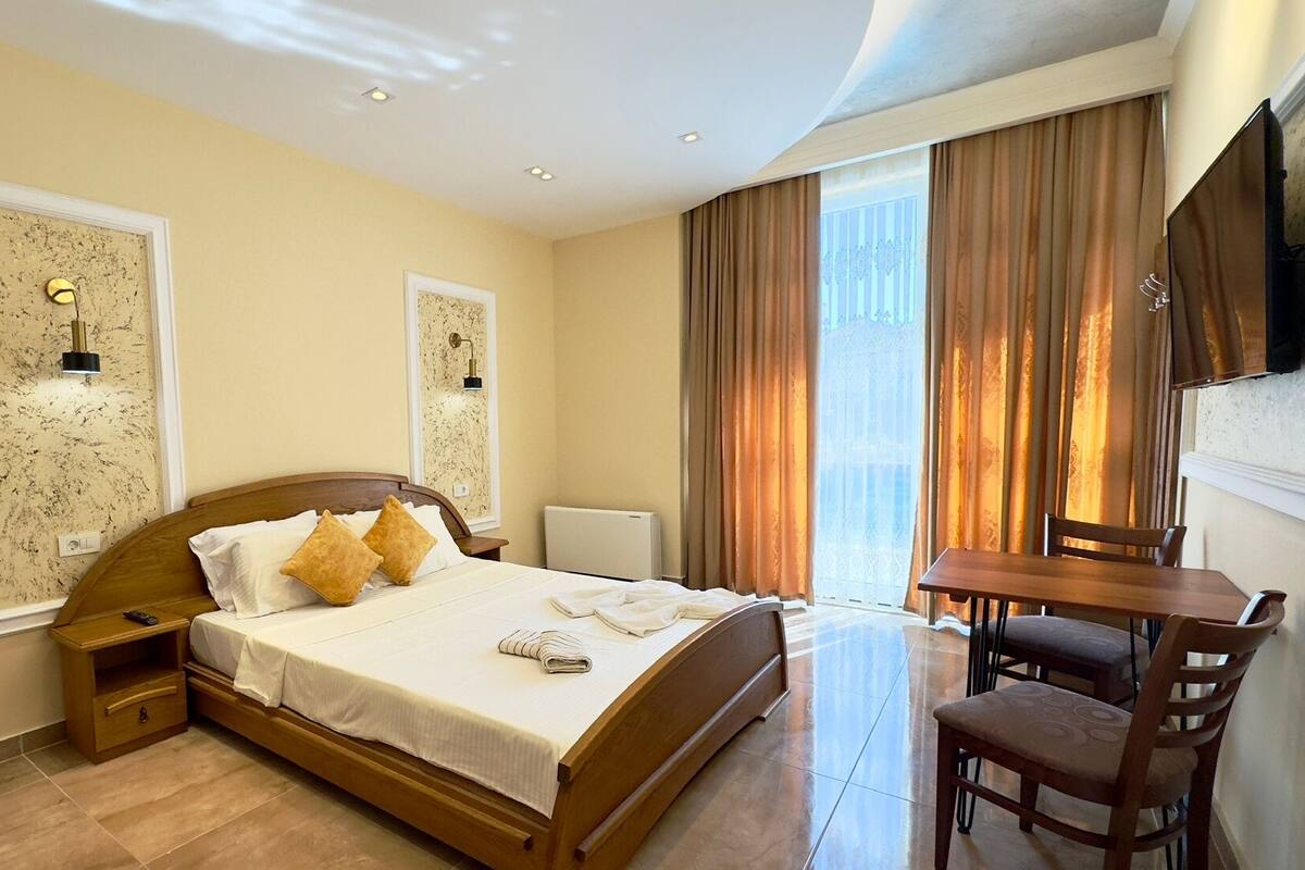 Ein Hotelzimmer mit einem Bett, einem Schreibtisch und einem Stuhl. Zwei Wandlampen und ein Fenster mit Vorhängen sind vorhanden.