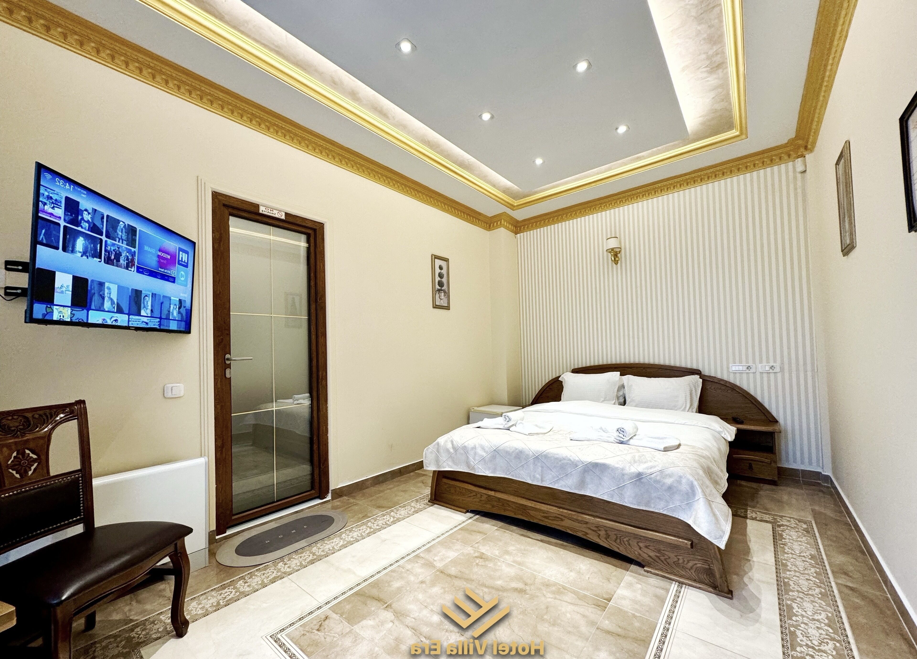 A hotel room with a bed, a TV mounted on the wall, a wooden chair, and a door leading to a bathroom.