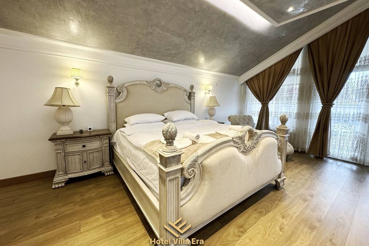 Ein geräumiges Schlafzimmer mit einem großen Bett, einem aufwendig verzierten Kopfteil und Blick auf die Stadt durch große Fenster.