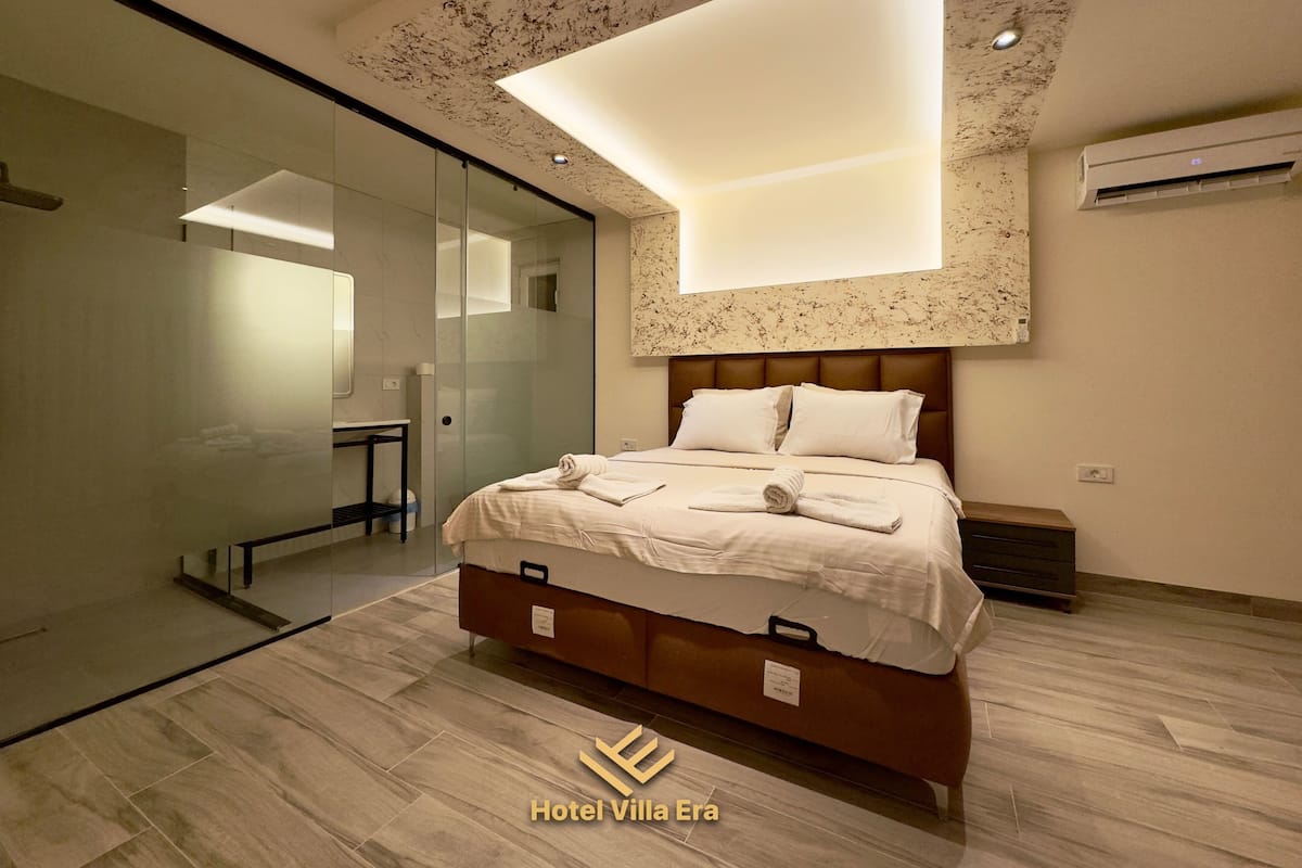 Ein modernes Hotelzimmer mit einer gläsernen Duschkabine, einem großen Bett mit weißen Bettwäsche und einer Deckenleuchte.