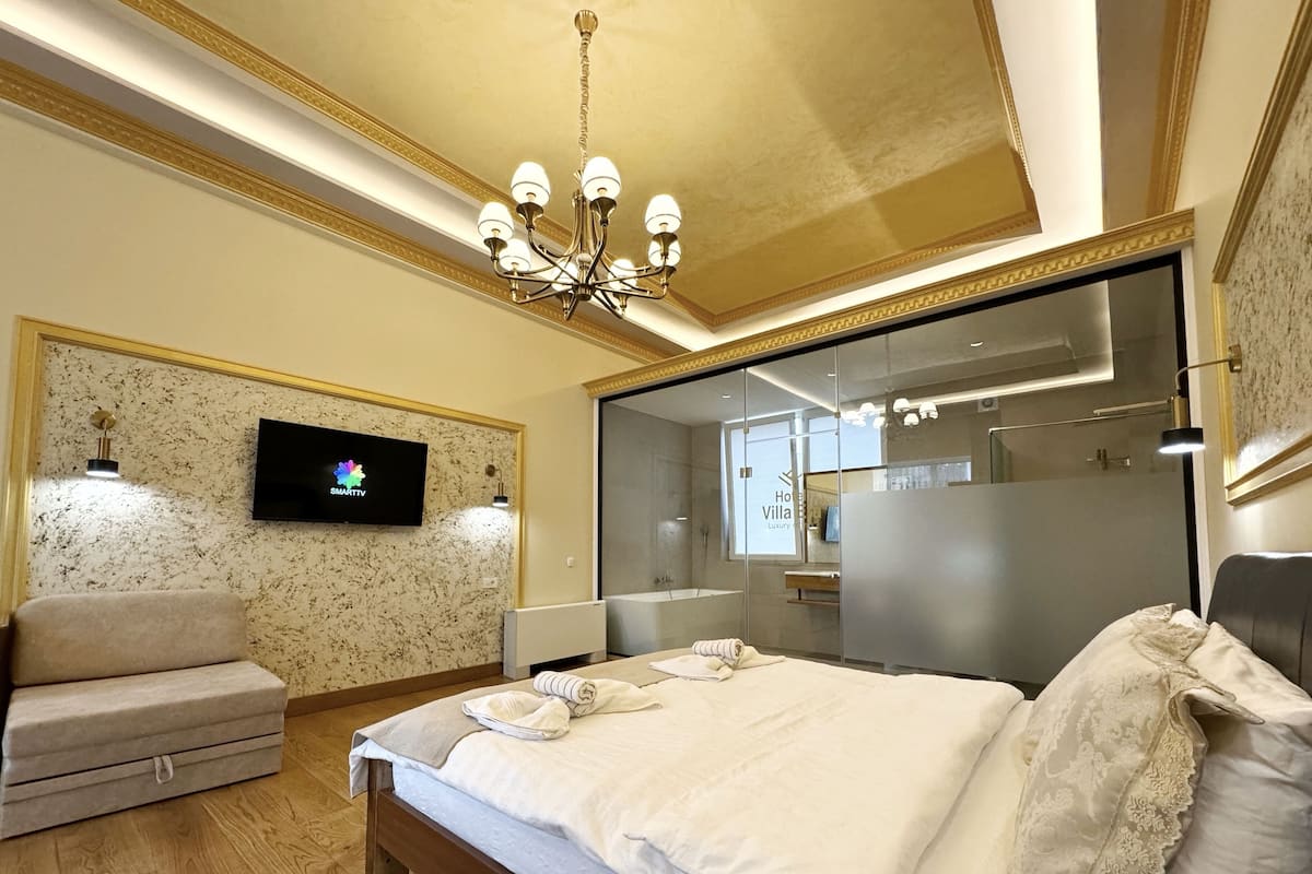 Ein modernes Hotelzimmer mit einem großen Bett, einem Flachbildfernseher, einer Couch und einem durch eine Glastrennwand einsehbaren Badezimmer.