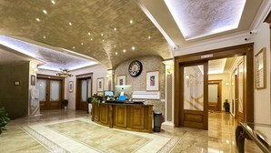 Vestíbulo de hotel con recepción, un reloj, cuadros enmarcados y una araña.