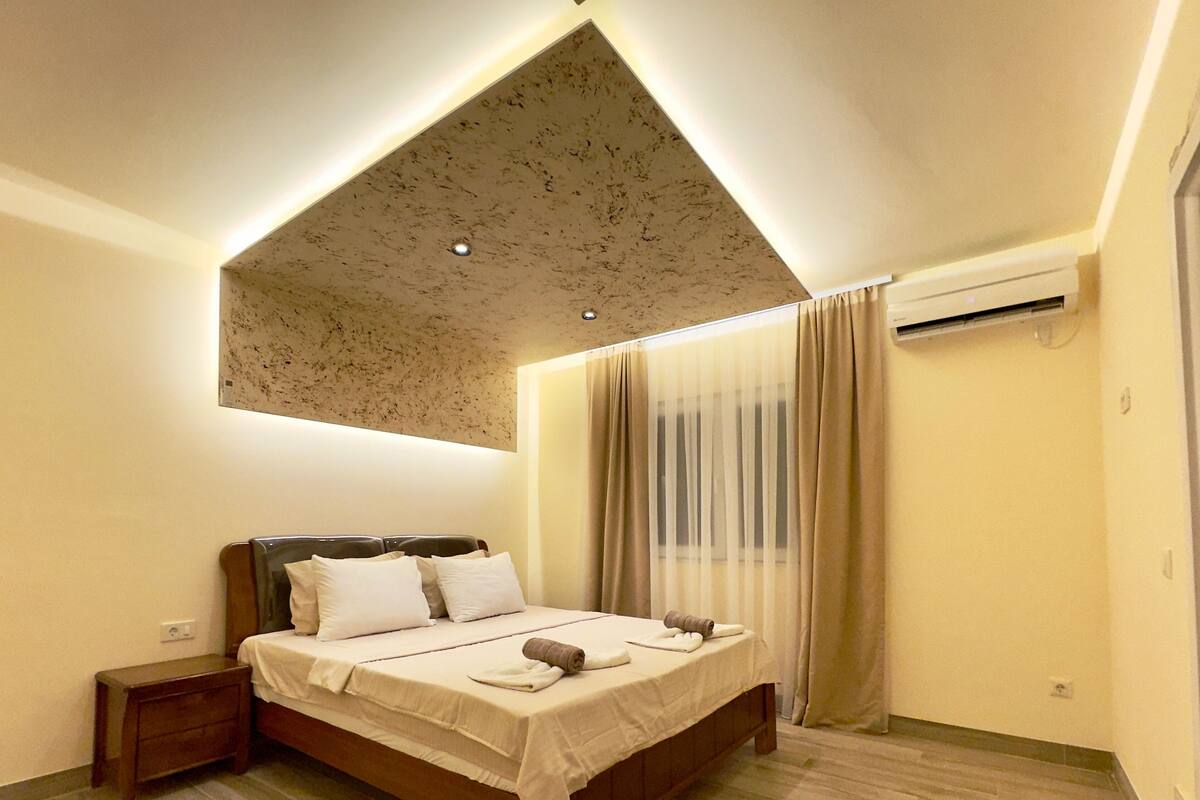 Ein Hotelzimmer mit Bett, Nachttisch, Deckenleuchte und einem Fenster mit Vorhängen.