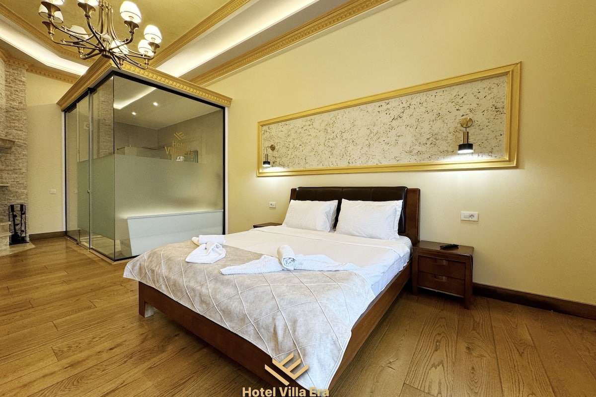 Ein Hotelzimmer mit einem großen Bett, einer Glasdusche, einer Chandelüstere und einem gerahmten Kunstwerk an der Wand.