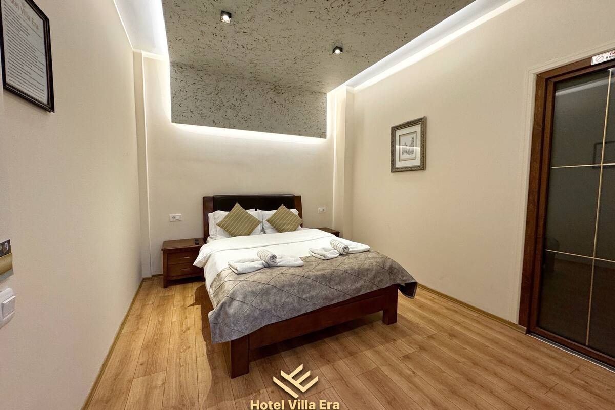 Ein modernes Hotelzimmer mit Holzboden, einem Bett mit grauer Steppdecke und einer Decke mit eingelassenen Leuchten.