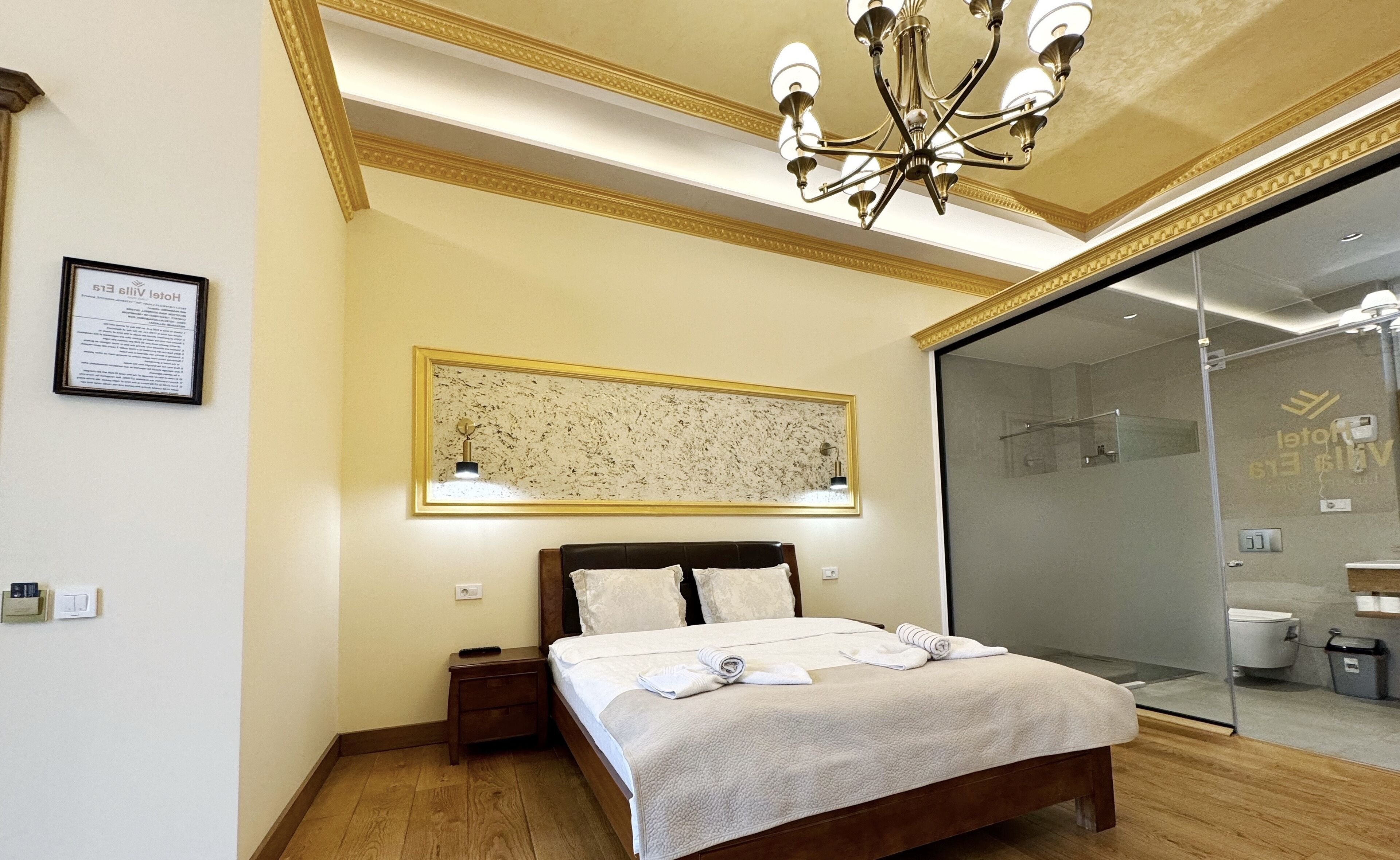 A hotel room with a bed, bedside table, nightstand, chandelier, wall-mounted artwork, and a glass shower enclosure.