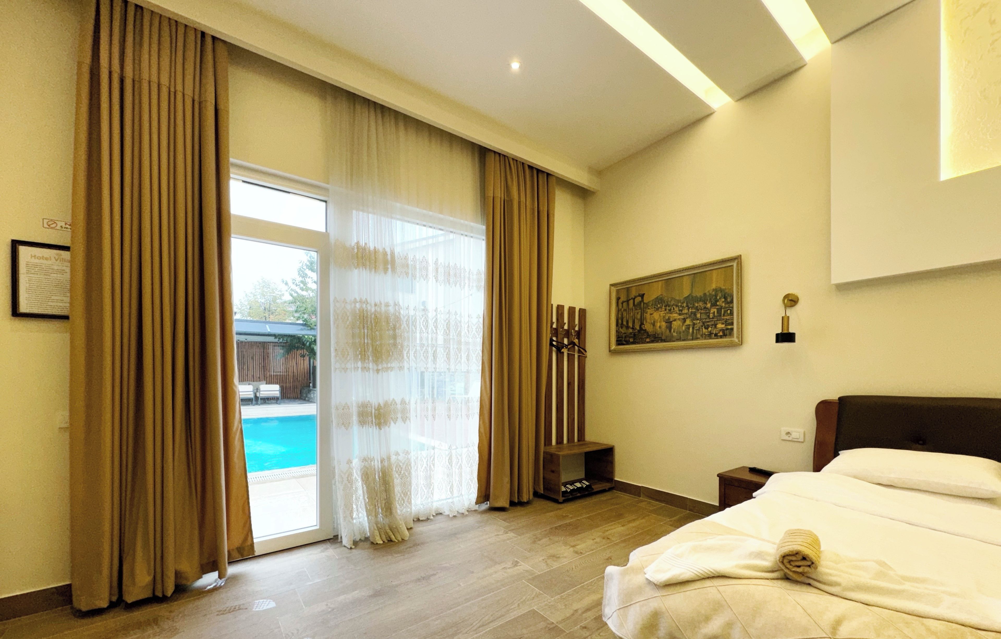 A hotel room with a bed, a painting on the wall, and a view of a swimming pool through the window.