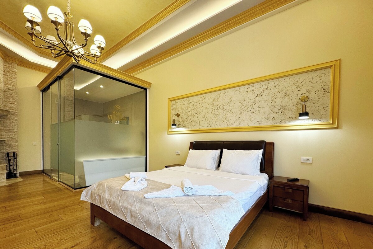 Habitación de hotel con una cama grande, cabina de ducha de vidrio, candelabro y una obra de arte enmarcada en la pared.