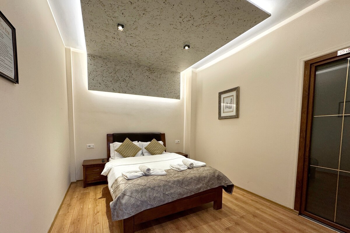 Una habitación de hotel moderna con piso de madera, cama con colcha gris e iluminación empotrada en el techo.