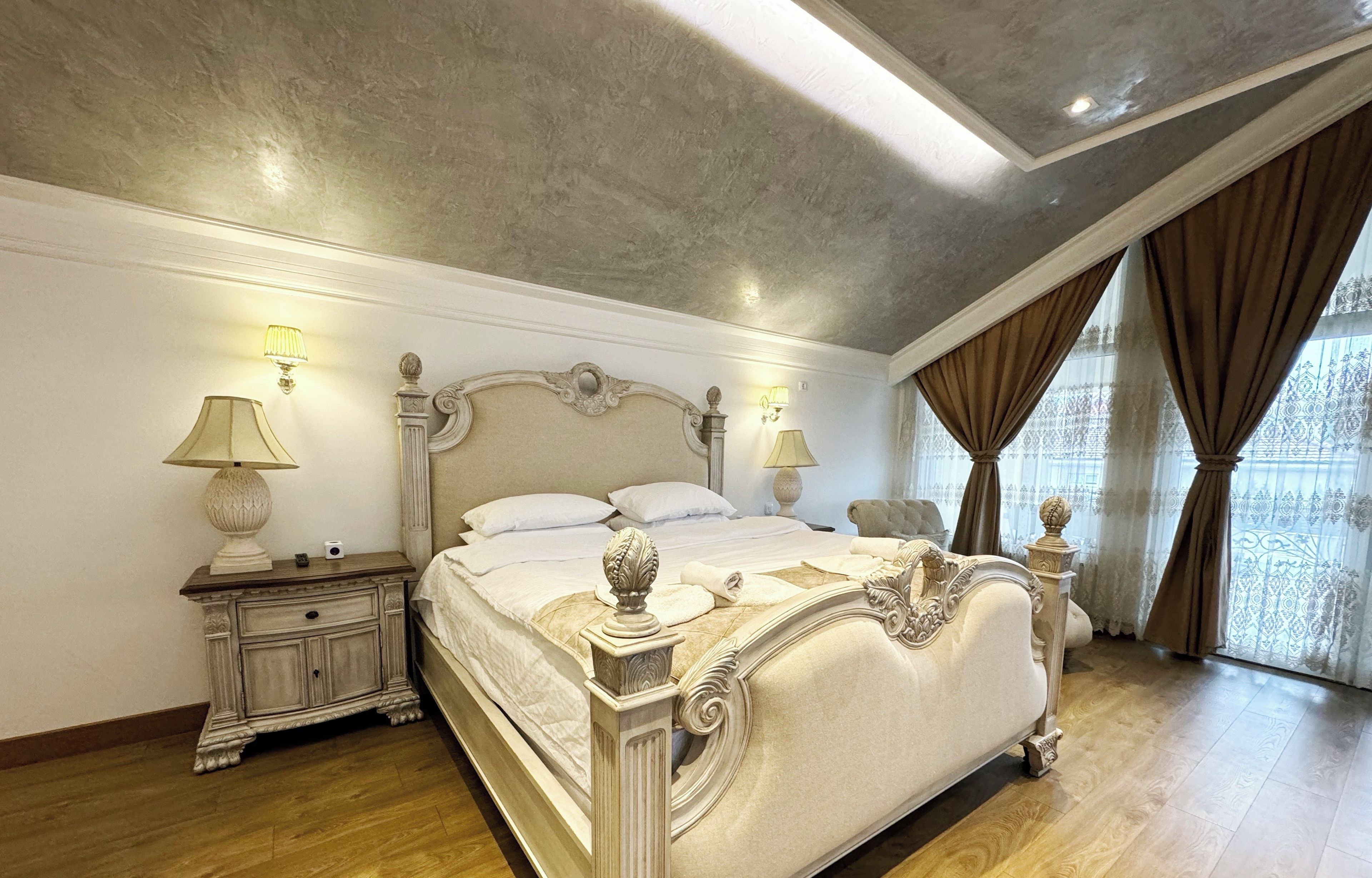 A spacious bedroom with a large bed, ornate headboard, and a view of the city through large windows.
