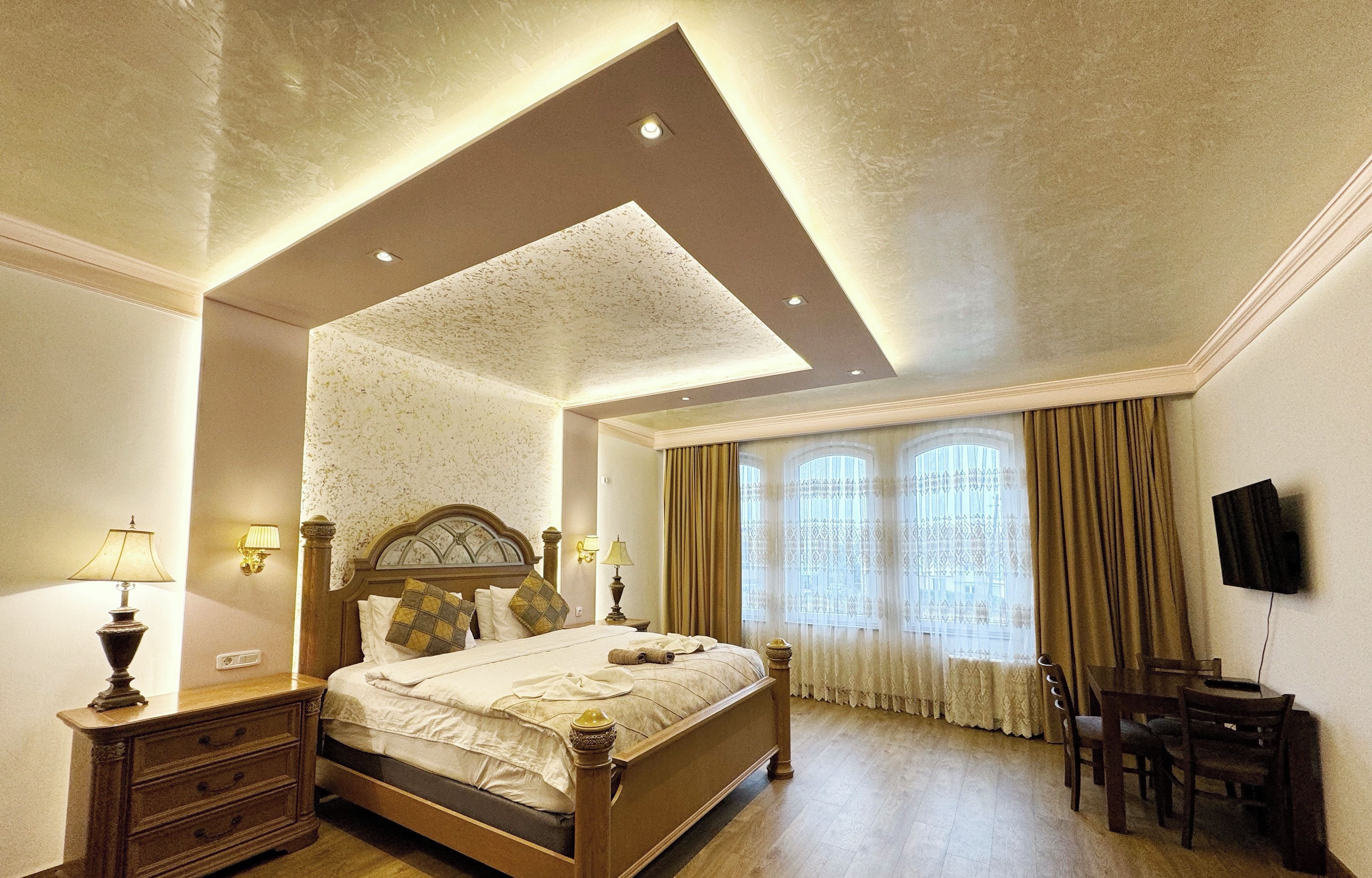 Una habitación de hotel con una cama grande, lámparas de noche, un aparador de madera, un escritorio con sillas y un televisor montado en la pared.