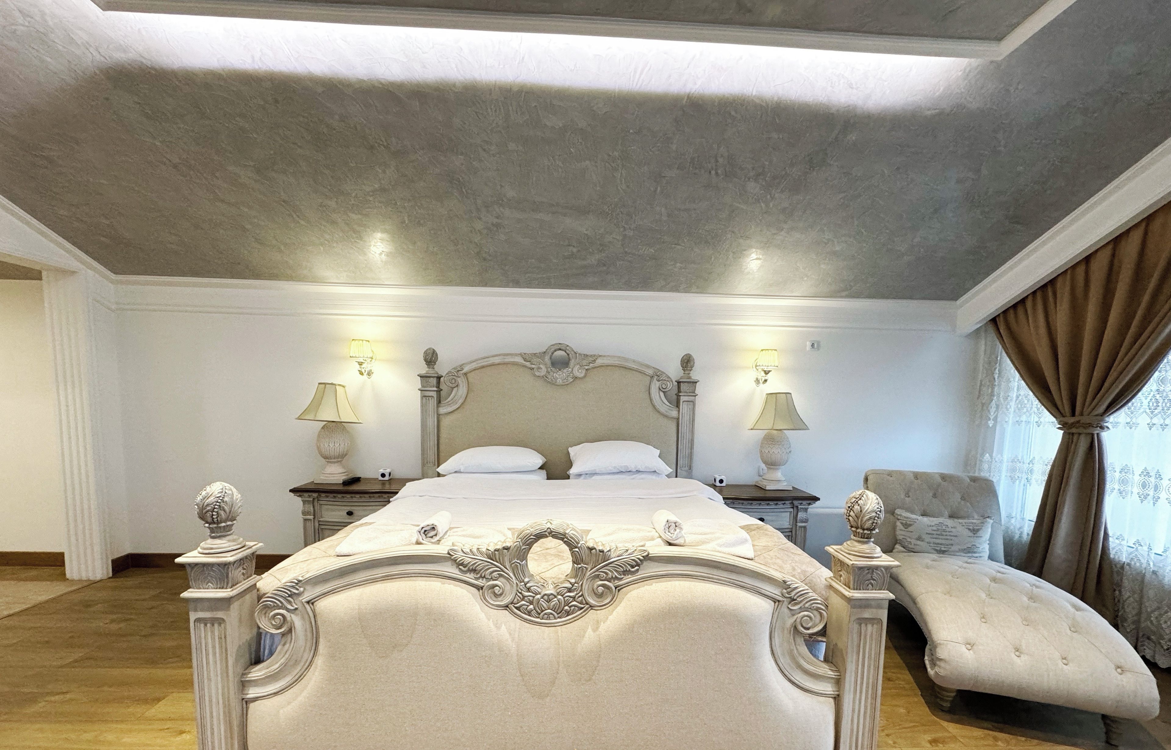 A spacious bedroom with a large bed, two bedside tables, a chaise lounge, and a ceiling light fixture.