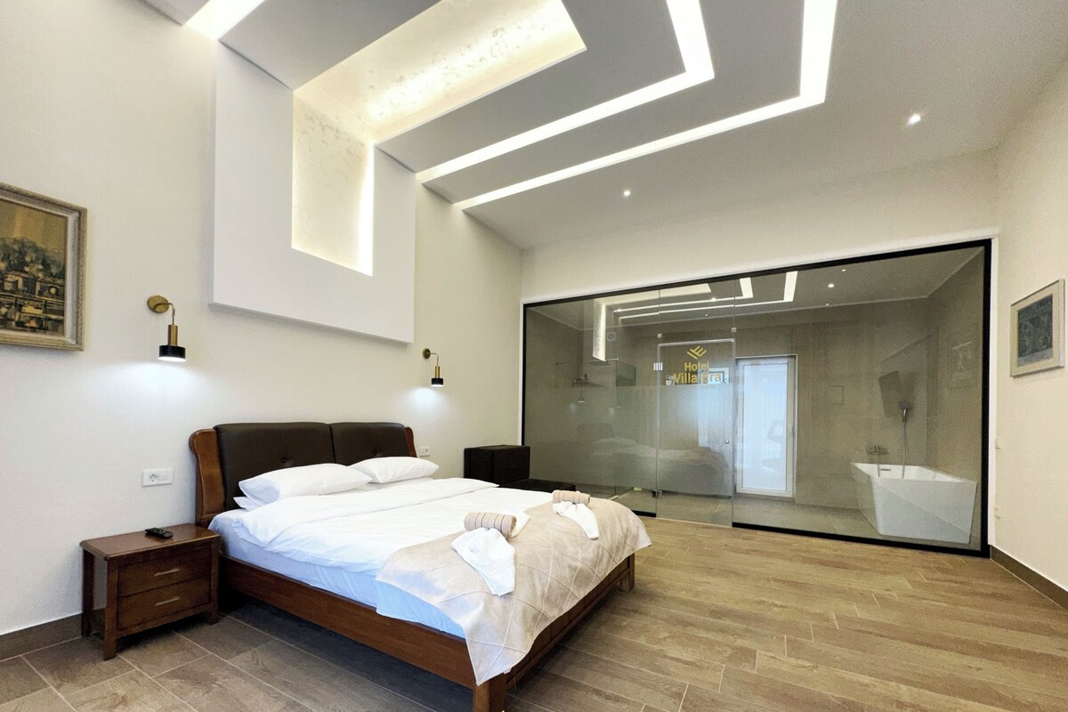 Habitación de hotel moderna con una cama grande, mesitas de noche, una lámpara de pared y un ventanal amplio.