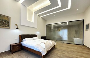 Habitación de hotel moderna con una cama grande, mesitas de noche, una lámpara de pared y un ventanal amplio.