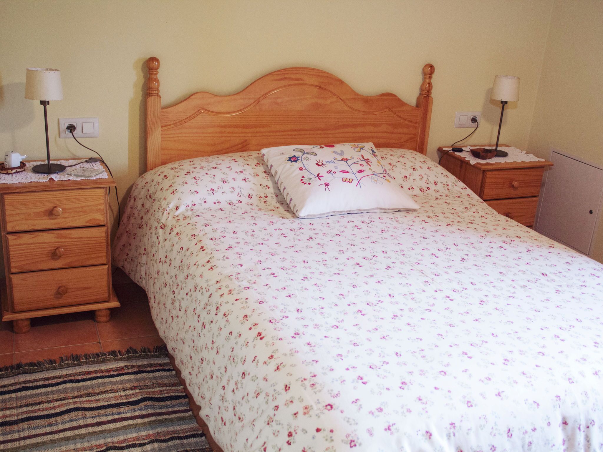 A bedroom with a wooden headboard, a floral bedspread, a decorative pillow, two bedside tables with lamps, and a small wooden dresser.