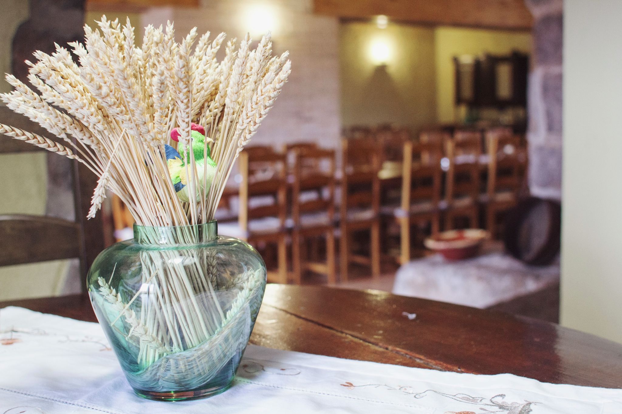 A glass vase with dried wheat stalks and a colorful chicken ornament on a table.