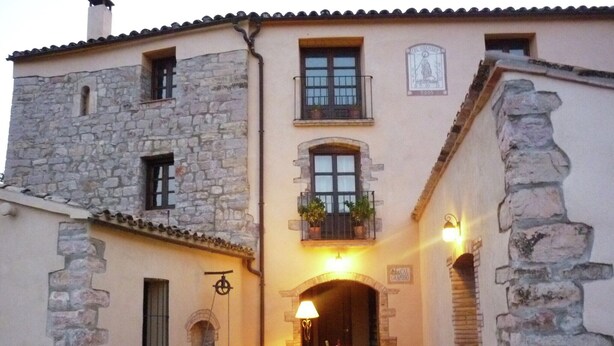 Un bâtiment traditionnel en pierre doté d’une cour, comprenant un espace repas avec des tables et des chaises, une cuve en pierre et un petit four en briques.
