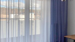 Una habitación con un ventanal grande cubierto por cortinas blancas translúcidas y una cortina azul opaca a la derecha.